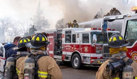 Firefighters in full gear are working at the scene of a fire. Several firefighters are facing a fire truck, with one standing on top, directing a hose at the burning building. Smoke is billowing in the background, and additional emergency vehicles are present.