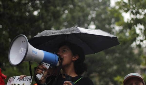 A woman is holding a megaphone and speaking into it while standing under an umbrella in the rain. She appears to be at an outdoor event or protest, with blurred greenery in the background.