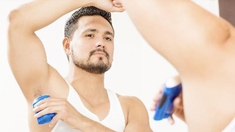 A man with a trimmed beard and short hair is applying deodorant to his underarm. He is looking at himself in the mirror and is wearing a white tank top.