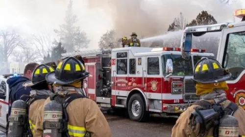Firefighters in full gear are working at the scene of a fire. Several firefighters are facing a fire truck, with one standing on top, directing a hose at the burning building. Smoke is billowing in the background, and additional emergency vehicles are present.