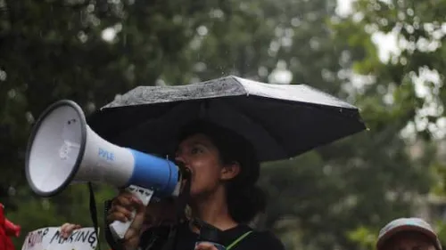 A woman is holding a megaphone and speaking into it while standing under an umbrella in the rain. She appears to be at an outdoor event or protest, with blurred greenery in the background.