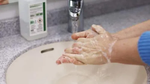 Hands covered in soap lather are being washed under running water in a sink. A bottle of liquid soap is visible on the counter next to the sink.