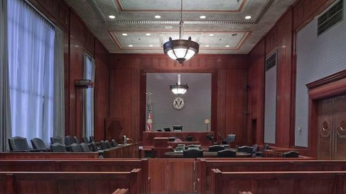 An empty courtroom featuring wooden furnishings, a judge's bench, jury seats, and a witness stand. The room is well-lit with chandeliers hanging from the ceiling, and an American flag is visible behind the judge's bench.