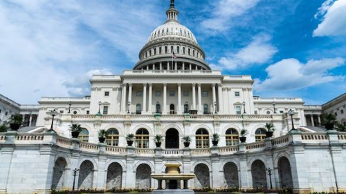 A view of the United States Capitol building in Washington, D.C., featuring its iconic dome and neoclassical architecture. The building is set against a blue sky with some clouds.