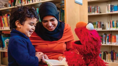 Photo of Afghan mother, child, and puppet from a television show smiling and reading a book in the library. Photo Credit: Sesame Workshop