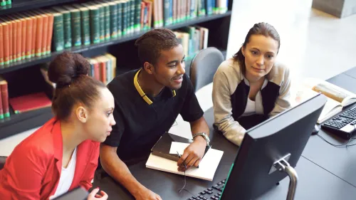 Three students look at a computer screen in a library with shelves of books in the background.