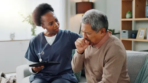 A healthcare provider offers support to a patient who is coughing into his hand while seated on a couch.