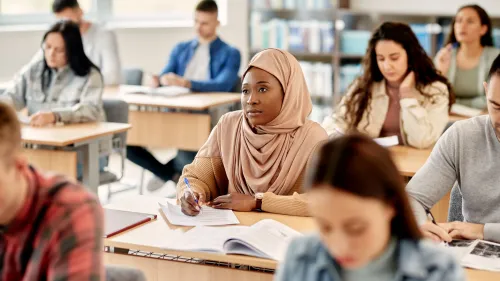 A female student wearing a hijab sits among other adult learners in a classroom.