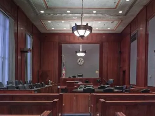 An empty courtroom featuring wooden furnishings, a judge's bench, jury seats, and a witness stand. The room is well-lit with chandeliers hanging from the ceiling, and an American flag is visible behind the judge's bench.