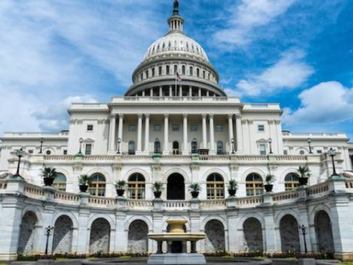 A view of the United States Capitol building in Washington, D.C., featuring its iconic dome and neoclassical architecture. The building is set against a blue sky with some clouds.