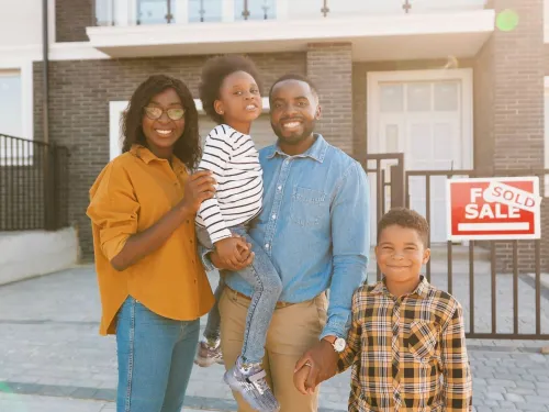Una familia de cuatro personas está parada frente a una casa que tiene un cartel de vendido en la puerta de entrada.