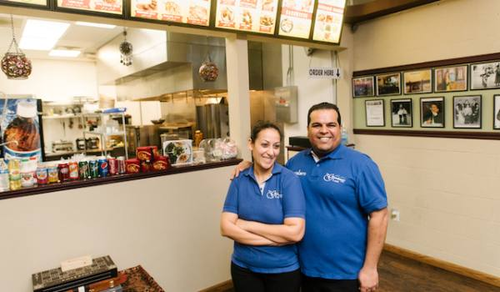 A man and a woman, both wearing blue polo shirts with the restaurant's name, are standing inside a restaurant. The woman has her arms crossed, and the man has his arm around her shoulder. Behind them is the counter with a menu board above, displaying various food items. The kitchen area and some framed pictures on the wall are also visible.