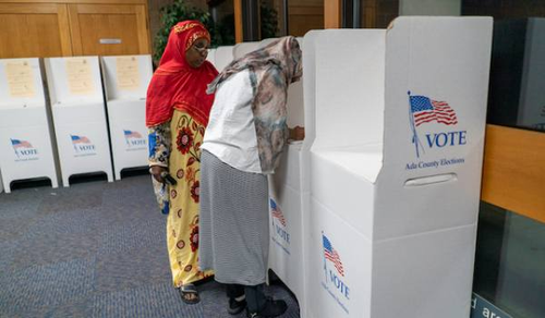 Two women in colorful traditional attire are voting at polling stations. One woman is wearing a red headscarf and yellow dress with a floral pattern, while the other woman is wearing a light-colored headscarf and grey dress. They are standing at white voting booths with the word 'VOTE' and an American flag printed on them.