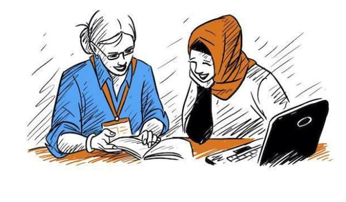 Two women are sitting at a table, engaged in a discussion. One woman, wearing glasses and a blue shirt with a lanyard, is pointing at a book. The other woman, wearing a hijab and a black dress, is attentively looking at the book. A laptop is open on the table beside them.