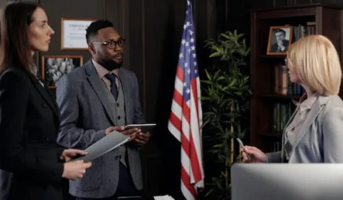 Three professionals are having a discussion in an office setting. A woman with long brown hair and a man with glasses and a beard, both holding tablets, are listening to a blonde woman who is speaking. An American flag stands in the background, along with bookshelves and framed photographs.