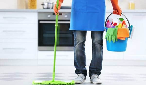 A person wearing jeans and an apron is holding a green mop in one hand and a blue bucket filled with cleaning supplies in the other hand. The background shows a clean kitchen with white cabinets and a stainless steel oven.