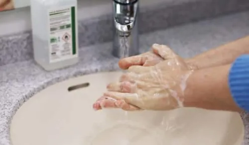 Hands covered in soap lather are being washed under running water in a sink. A bottle of liquid soap is visible on the counter next to the sink.
