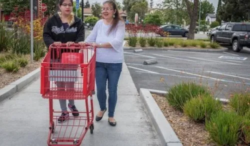 Two women are walking on a sidewalk in a parking lot, pushing a red shopping cart. One woman is wearing a black hoodie, and the other is wearing a white blouse. They appear to be having a conversation as they walk.