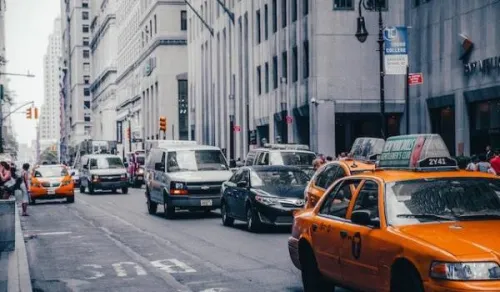 A busy city street with multiple lanes of traffic, including yellow taxis, vans, and cars. Tall buildings line both sides of the street, and pedestrians are visible on the sidewalks. The scene captures the hustle and bustle of urban life.