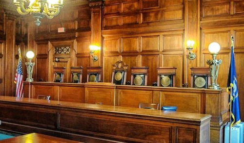 An empty courtroom with a large wooden bench and five high-backed chairs for judges. The room is decorated with wood paneling and lit by ornate lamps. An American flag and another flag stand to the side.