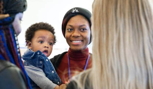 A smiling woman wearing a black hat is holding a baby and standing in a group with other women. The baby is looking curiously at one of the women in the group. The setting appears to be casual and friendly.