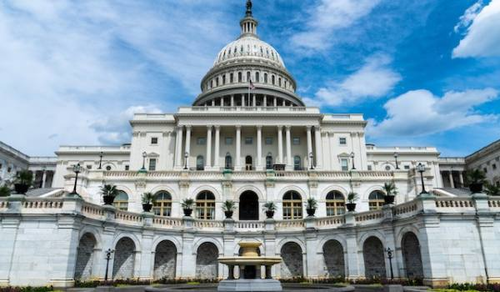 A view of the United States Capitol building in Washington, D.C., featuring its iconic dome and neoclassical architecture. The building is set against a blue sky with some clouds.