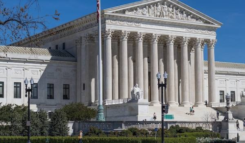 A view of the United States Supreme Court building, featuring its grand neoclassical architecture with tall columns and a detailed pediment. An American flag is flying in front of the building, and the scene is set against a clear blue sky.