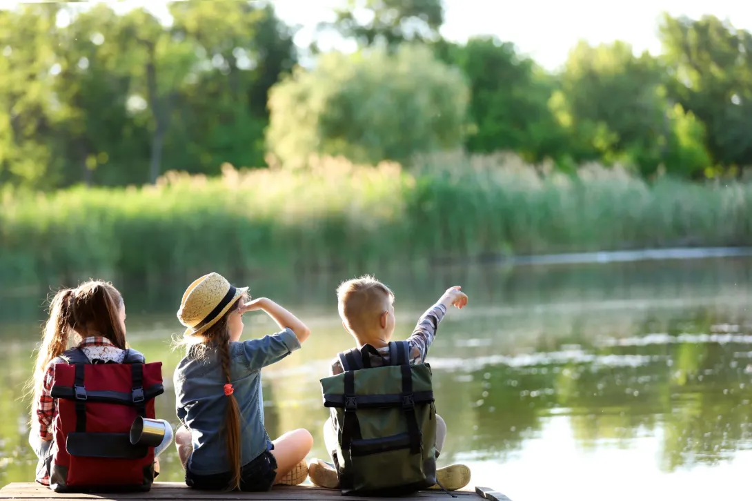 Three children sitting on a dock next to a body of water. One child is pointing across the water.