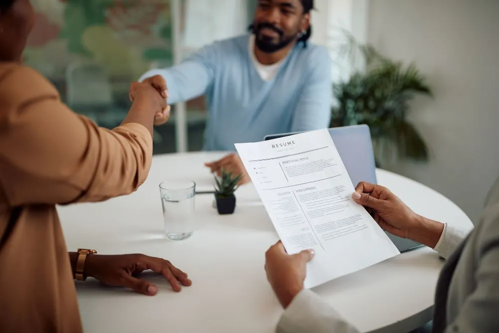 Two people shaking hands at a job interview. One person is reading a resume.