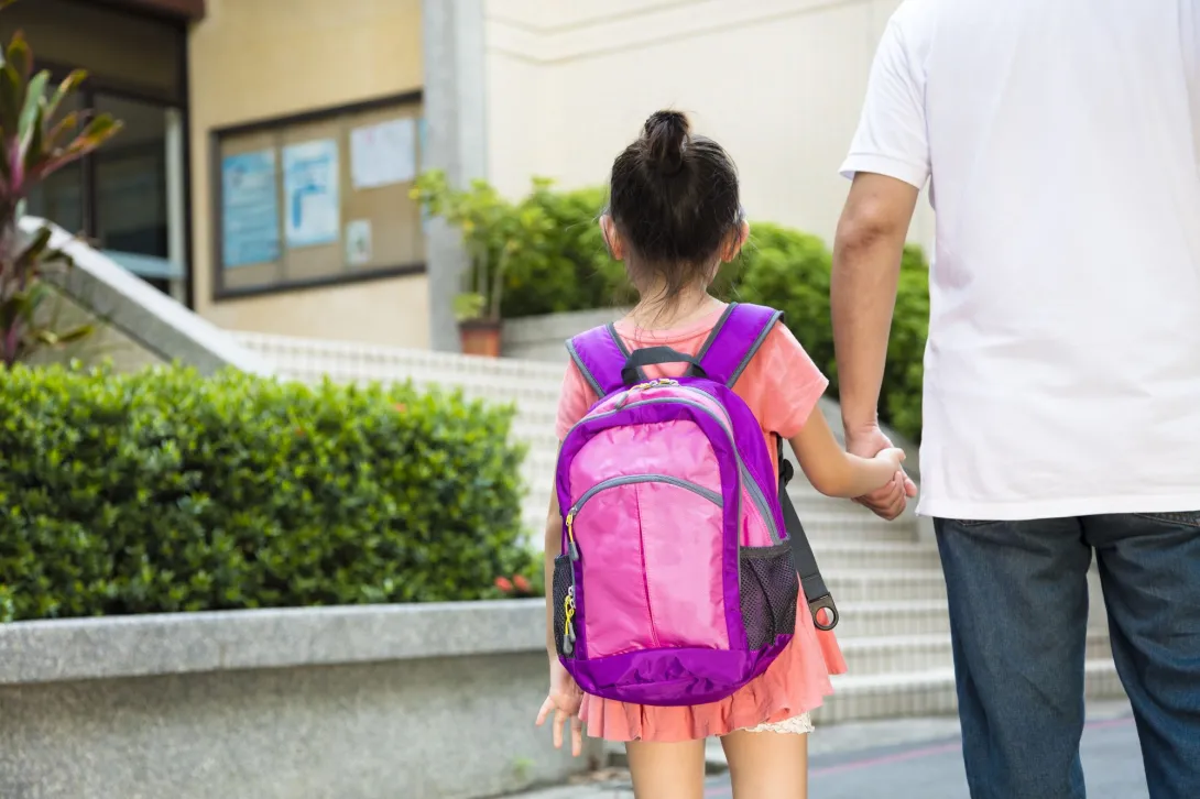 An adult is holding their child’s hand as they walk into the school building. The child is wearing a backpack.